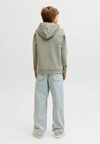 Pilkas-žalias džemperis su laisvu siluetu, turintis gobtuvą ir rišančius rankogalius, derinamas su šviesiai mėlynomis plačiomis kelnėmis ir neutralios spalvos batais.