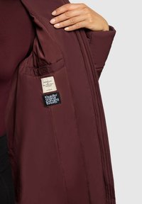 Burgunderfarbene gepolsterte Jacke mit glatter Textur, mit einem Etikett und einer Tasche im Innenfutter. Doppelte Nähte entlang der Nähte.