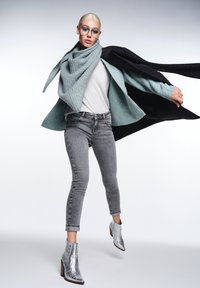 En person bär en ljus teal-färgad stickad halsduk, grå överdel, grå jeans med utsmyckningar och silverfärgade klackboots mot en neutral bakgrund.