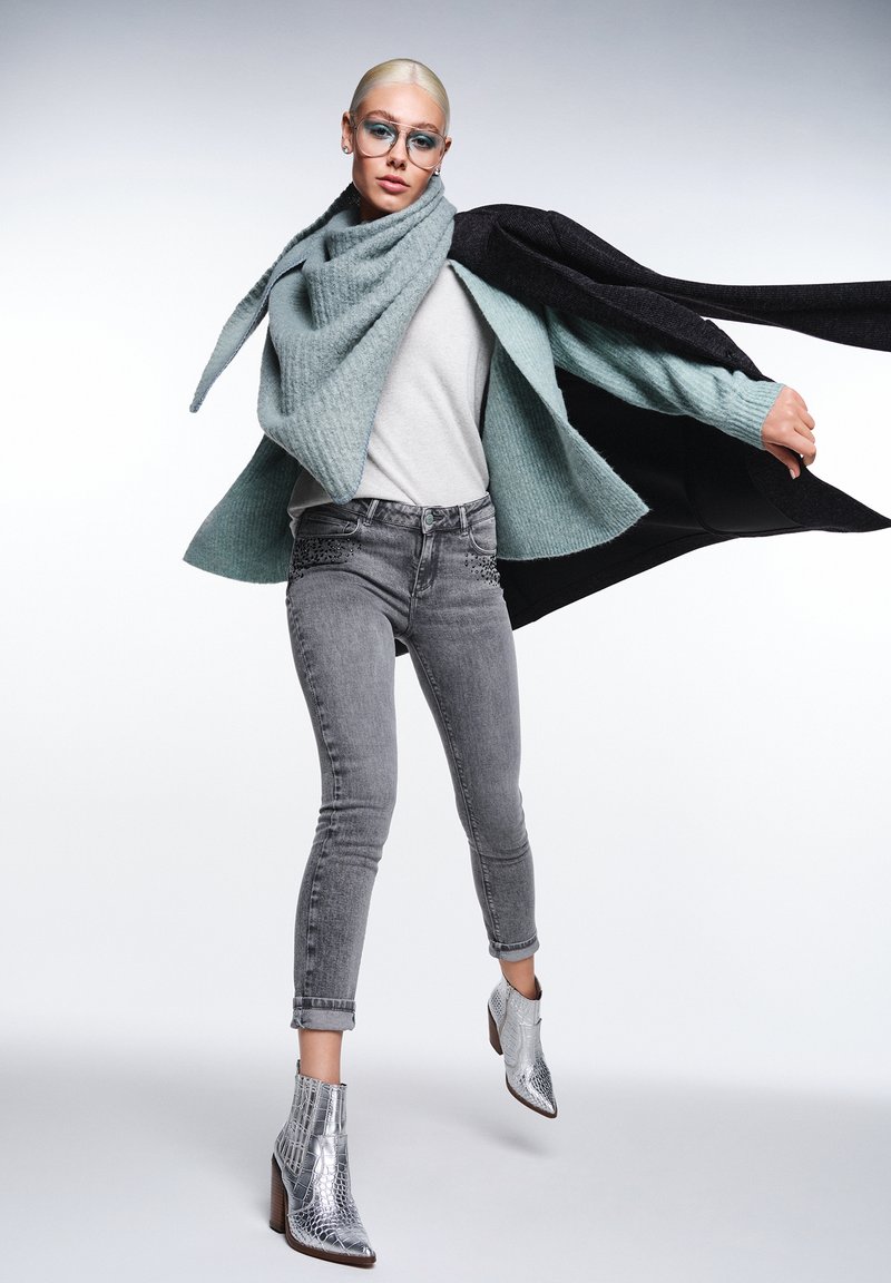 En person bär en ljus teal-färgad stickad halsduk, grå överdel, grå jeans med utsmyckningar och silverfärgade klackboots mot en neutral bakgrund.