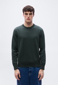 Νέο ς άνδρας που φοράει σκούρο πράσινο πουλόβερ με στρογγυλή λαιμόκοψη και λογότυπο "BOSS" και μπλε τζιν, στέκεται μπροστά σε απλό λευκό φόντο.