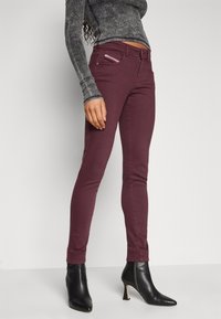 Jeans burdeos ajustados con una cinturilla alta, bolsillos con cremallera y una textura suave, combinados con estiletos negros de punta.