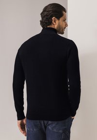 Schwarzer Strickpullover mit hohem Kragen, der eine gerippte Textur und lange Ärmel aufweist, von hinten vor neutralem Hintergrund gesehen.