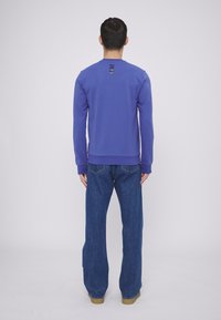 Felpa viola con maniche lunghe e scollo rotondo. Presenta un accento sul retro con tagli orizzontali. Abbinata a jeans blu a gamba larga.