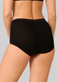 Donna che indossa mutandine di cotone nere a vita alta con pizzo, vista da dietro su uno sfondo grigio uniforme.