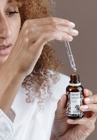 Brun glassflaske med blemish serum og hvit, svart og rosa etikett; pipette dispenserer klar væske, holdt av en hånd med lys hudtone.