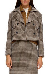 Skórzana kurtka w kratę w odcieniach brązu i beżu, z dużymi klapami, podwójnym zapięciem na guziki i dwoma przednimi kieszeniami. Nałożona na brązową bluzę z zamkiem.