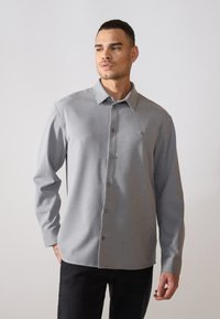 Camicia grigia con bottoni, a maniche lunghe, realizzata in un tessuto testurizzato, con colletto classico e un piccolo emblema ricamato sul petto.