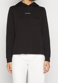Persona che indossa una felpa nera Calvin Klein con il logo bianco centrato sul petto e pantaloni bianchi, in piedi davanti a uno sfondo neutro.