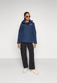 Marineblauw waterdicht jack met capuchon, ritssluiting en logo op de borst, gecombineerd met zwarte cargobroek en beige schoenen. Verstelbare manchetten en zakken.