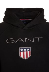 Czarna bluza z kapturem z logo "GANT" w kolorze szarym, z emblematem tarczy przedstawiającym czerwone i białe paski oraz trzy białe gwiazdy. Miękka faktura tkaniny.