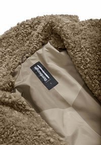 Close-up van een beige krullende nepbontjas met een gladde binnenvoering, voorzien van een zwart BomBoogie merketiket en maat 2 label.