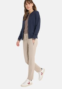 Námořnický kardigan přes hnědé tričko, béžové tepláky s bočními kapsami a bílé tenisky s červenými detaily. Stylové vrstvení pro ležérní nošení.