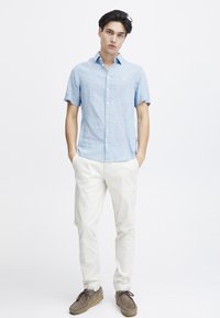 Camicia azzurro chiaro a maniche corte con bottoni realizzata in lino, abbinata a pantaloni bianchi e scarpe grigie. Presenta una texture sottile e un logo.