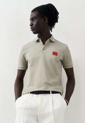 Man with dreadlocks wearing a beige polo shirt with a red Hugo logo patch and white pants, hands in pockets, looking to the side.