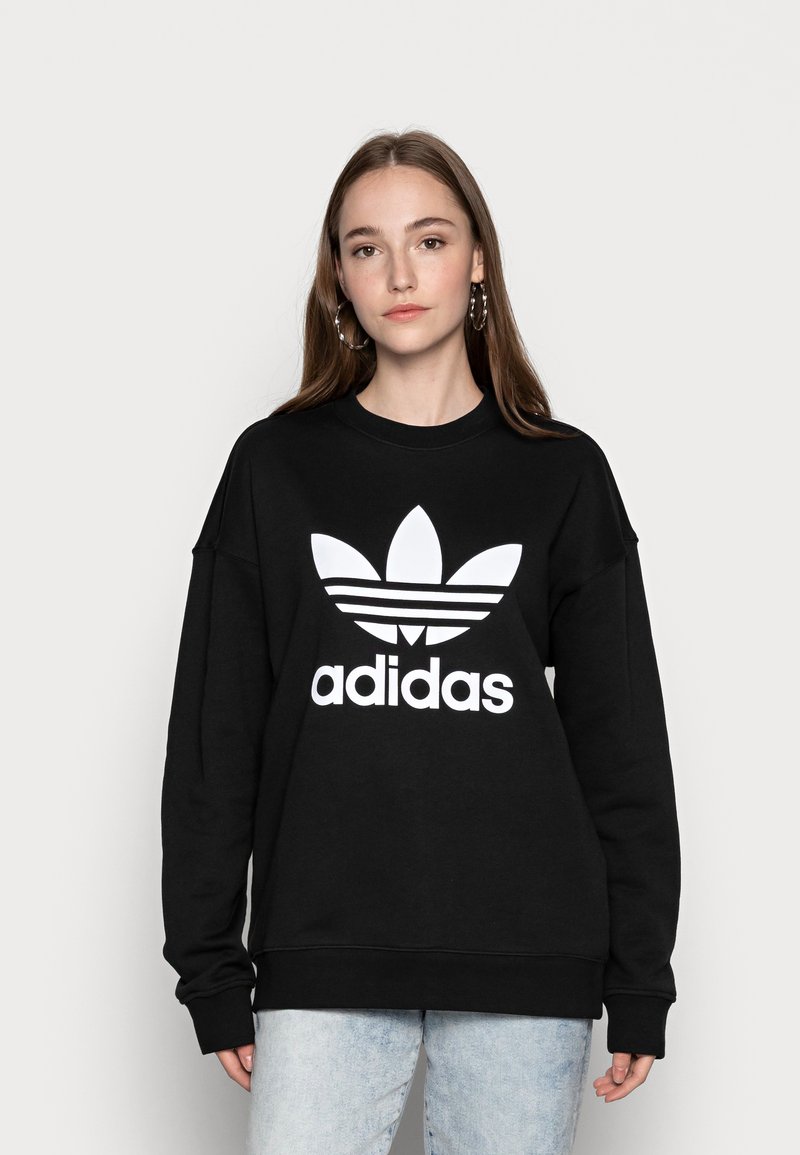 Crna oversized dukserica od pamuka, s velikim bijelim Adidas logotipom i trefoy dizajnom u središtu. Jednostavan, ležeran stil.
