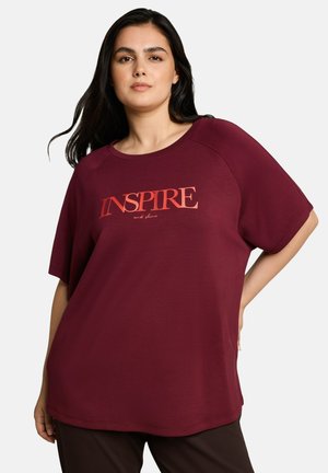 Frau mit langen dunklen Haaren trägt ein locker sitzendes weinrotes T-Shirt mit "INSPIRE and shine", gedruckt in roten und orangen Buchstaben auf hellem Hintergrund.