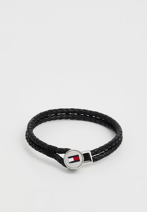 Brăță din piele neagră împletită, cu un închizător din argint care prezintă un logo circular cu detalii roșii și albastre. Textură netedă, design cu două curele.