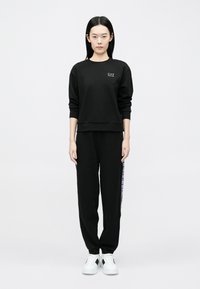 Žena stojaca, oblečená v čiernej mikine s logom "EA7" a čiernych teplákoch s bočnými pásmi, biele tenisky, na bielom pozadí.