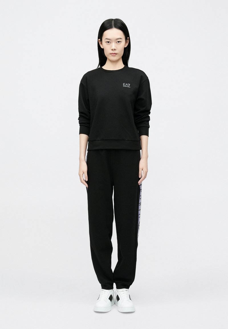 Žena stojaca, oblečená v čiernej mikine s logom "EA7" a čiernych teplákoch s bočnými pásmi, biele tenisky, na bielom pozadí.