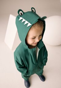 Grøn vandtæt heldragt med hætte, der har øjne og tænder, lynlåslukning og elastiske manchetter. Glat tekstur, afslappet design.