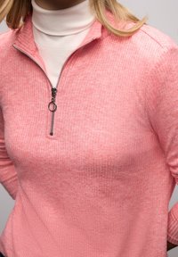 Vaaleanpunainen neljäsosa-vetoketjullinen pusero, jossa on resorikuvio ja hopeinen vetoketju. Päälle puettu valkoinen poolopaita luo kontrastisen ilmeen.