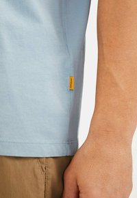 Lyseblå T-shirt lavet af bomuld, med et logo tag i siden i klart orange, korte ærmer og en rund kant.