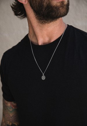 Colgante de plata en una cadena delgada, con un diseño circular y detalles intrincados, llevado sobre una camiseta negra.