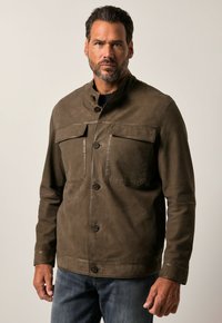 Bruine leren jas met een staande kraag, twee borstzakken, knoopsluiting en een gestructureerde afwerking. Gemaakt over een zwart top.