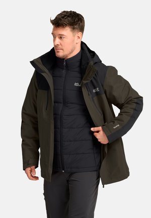 Uomo con giacca Jack Wolfskin nera e verde scuro, che sistema la cerniera frontale, rivolto verso sinistra su sfondo semplice.