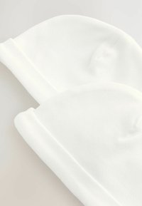 Dwie miękkie białe bawełniane czapeczki dla niemowląt z zawiniętymi brzegami ułożone jedna na drugiej na jasnym tle.