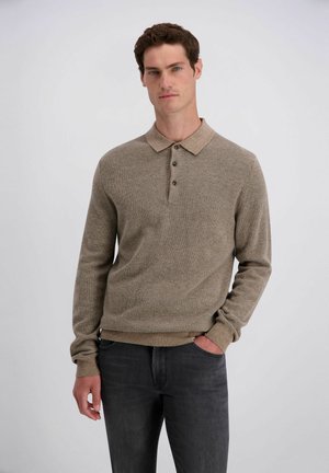 Man die een bruin poloshirt met lange mouwen en een donkere spijkerbroek draagt en met één hand in de zak staat tegen een effen lichte achtergrond.