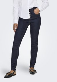 Donkerblauwe skinny jeans met hoge taille, voorzien van een vijfknops sluiting en een voorkantzak, aangevuld met contrasterende stikseldetails.