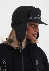 Musta pehmendatud müts, millel on kõrvaflapid, jäik sääk ja valge tikand esiosas. Sile tekstuur, reguleeritavad külgklambrid.