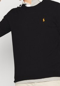 Svart tröja med gult broderat hästlogo, bärs över en vit skjorta med krage och en ljusgrå t-shirt under, tillsammans med beige byxor.