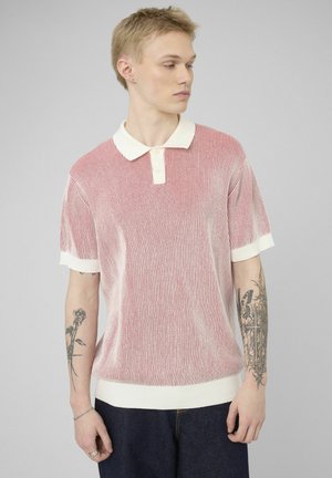 Jeune homme portant un polo à manches courtes rayé rouge et blanc avec des tatouages sur les deux avant-bras, debout devant un fond gris uni.