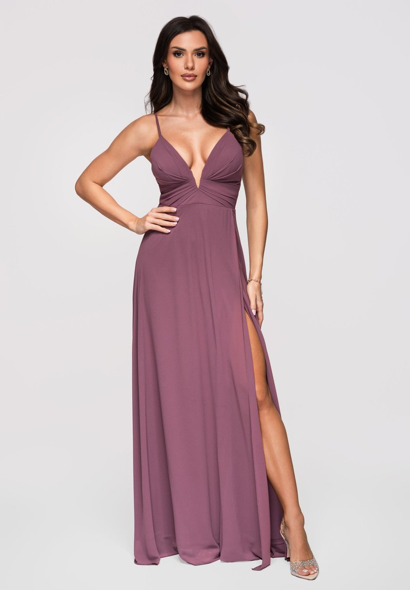 Femme aux longs cheveux foncés portant une robe mauve sans manches avec un décolleté en V profond et une fente haute, posant avec une main sur la hanche.