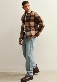 Camicia di flanella a scacchi in marrone, beige e blu, indossata sopra una t-shirt bianca, abbinata a jeans azzurri chiari e stivaletti marroni.