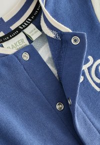Blaue Baumwolljacke mit geripptem Kragen, Druckknöpfen und gesticktem Text. Verfügt über einen strukturierten Stoff mit einem gestreiften Innenfutter.