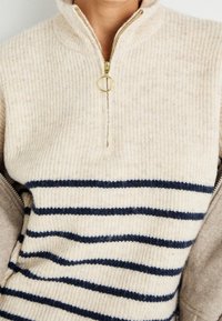 Suéter beige de media cremallera con rayas horizontales en azul marino, textura acanalada y herrajes de cremallera dorados. Tejido suave y de punto con un diseño casual.