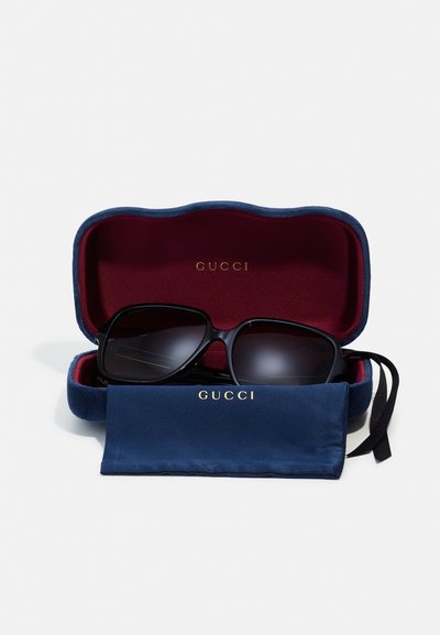 Sluneční brýle Gucci uvnitř námořnicky modré sametové krabičky s burgundským interiérem. Brýle mají tmavé čočky a černé obroučky.