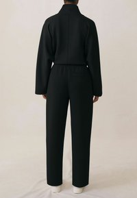 Czarny kombinezon z wysokim kołnierzem, długimi rękawami, elastycznym pasem i boczną kieszenią. Gładki materiał z uporządkowanym fasonem i zwężającymi się nogawkami.