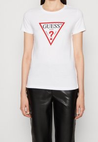 Camiseta de algodón blanca con un logo triangular rojo que lleva la palabra "GUESS" y un "?". Mangas cortas, cuello redondo, combinada con pantalones de cuero sintético negros.
