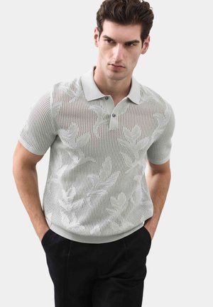 Mand iført lysegrå strikket kortærmet poloshirt med bladmønstre og sorte bukser, stående med hænderne i lommerne mod en ensfarvet baggrund.