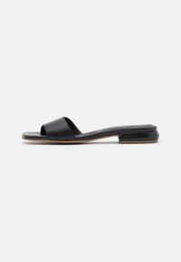 Sandalia deslizante de cuero negro con una tira ancha y un bajo tacón cuadrado. Presenta una textura suave y un diseño minimalista. Silueta de punta abierta.