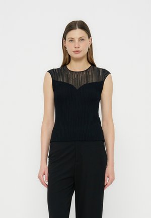 Γυναίκα φοράει μαύρο αμάνικο τοπ με διαφανή ριγέ στάγκλ και μαύρο παντελόνι, στέκεται μπροστά σε απλό λευκό φόντο.