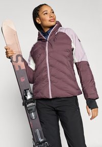 Vijolična in svetlo vijolična podložena jakna s kapuco, združena s črnimi smučarskimi hlačami in rožnatimi smučmi s črnimi vezmi na njih.