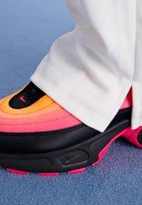 Nike tenisice s gradijentom narančaste i ružičaste boje, crnim akcentima i teksturiranom površinom, u kombinaciji s bijelim hlačama.