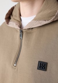 Bluza z kapturem w beżowym materiale z przednim zamkiem błyskawicznym, z teksturowanym beżowo-kremowym kapturem i czarnym kwadratowym logo na piersi.