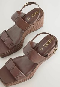 Brown wedge sandals with two wide straps, featuring decorative stitching, a textured heel, and a gold buckle on the ankle strap.
